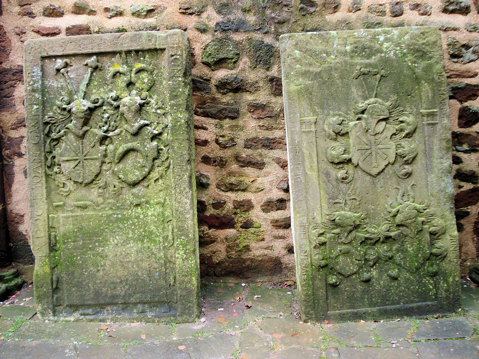 Zwei aufrecht stehende Wappengrabsteine an einer Bruchsteinmauer der Totenkirche.