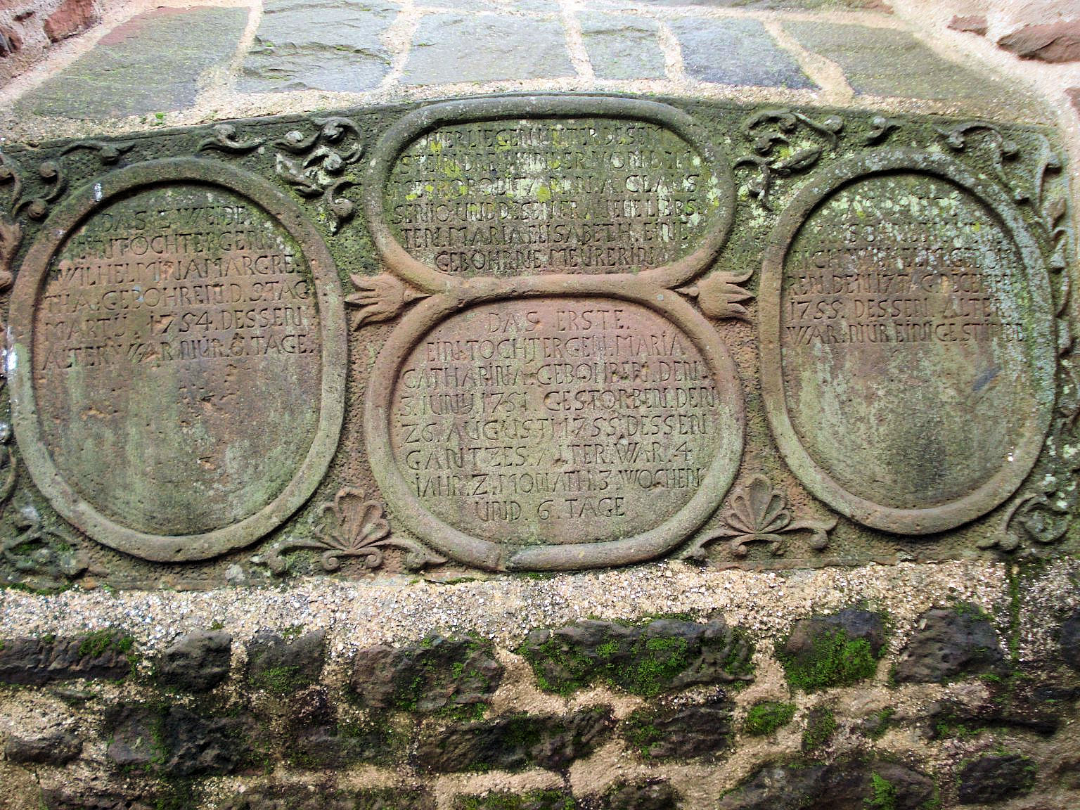 Sammel-Gedenkplatte mit drei ovalen Schriftfeldern in floraler Sandsteinrahmung, eingelassen in das Mauerwerk.